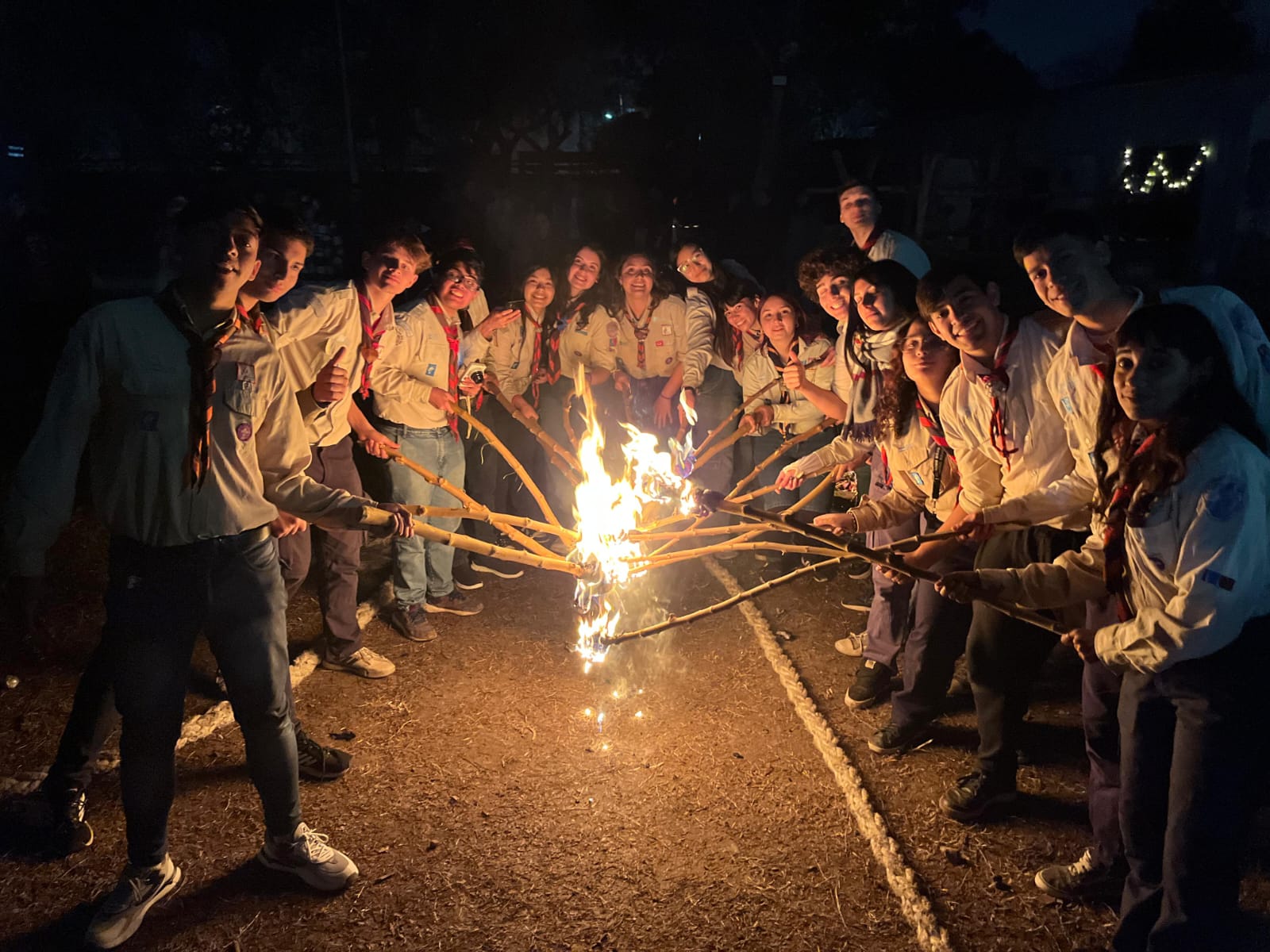 Jóvenes scouts reunidos alrededor de un fogón nocturno, sosteniendo antorchas encendidas en una actividad grupal, reflejando el compañerismo, la tradición y el espíritu scout.